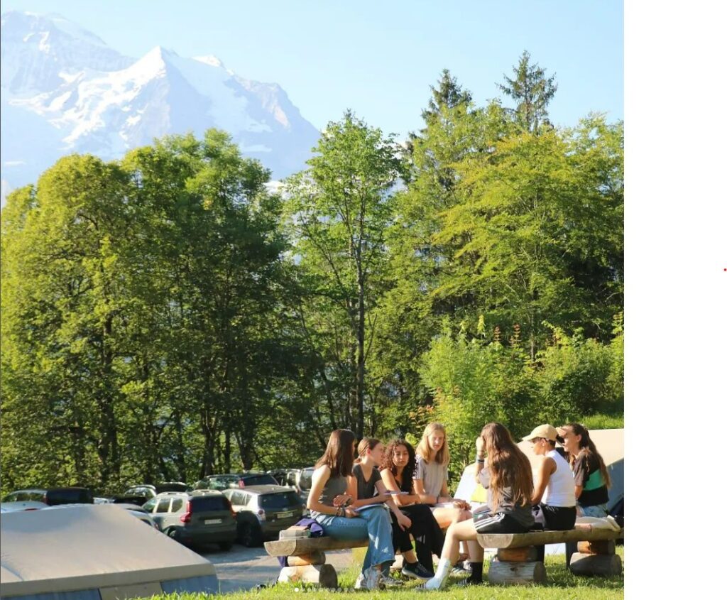 summer youth camp with group of people sitting on outdoor camp benches, there is a mountain in the distance, trees , and the tents