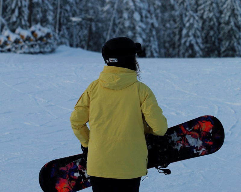 fille prise de dos , avec veste jaune qui porte son surf suite À une descente de piste